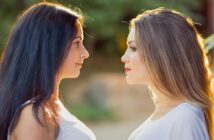 Two women with long hair are facing each other outdoors. One has dark hair and the other has light hair. Both are wearing white tops, and the background is softly blurred with greenery.