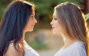 Two women with long hair are facing each other outdoors. One has dark hair and the other has light hair. Both are wearing white tops, and the background is softly blurred with greenery.