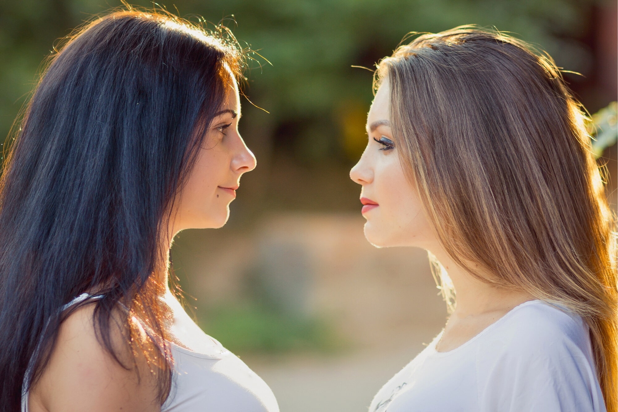 Two women with long hair are facing each other outdoors. One has dark hair and the other has light hair. Both are wearing white tops, and the background is softly blurred with greenery.