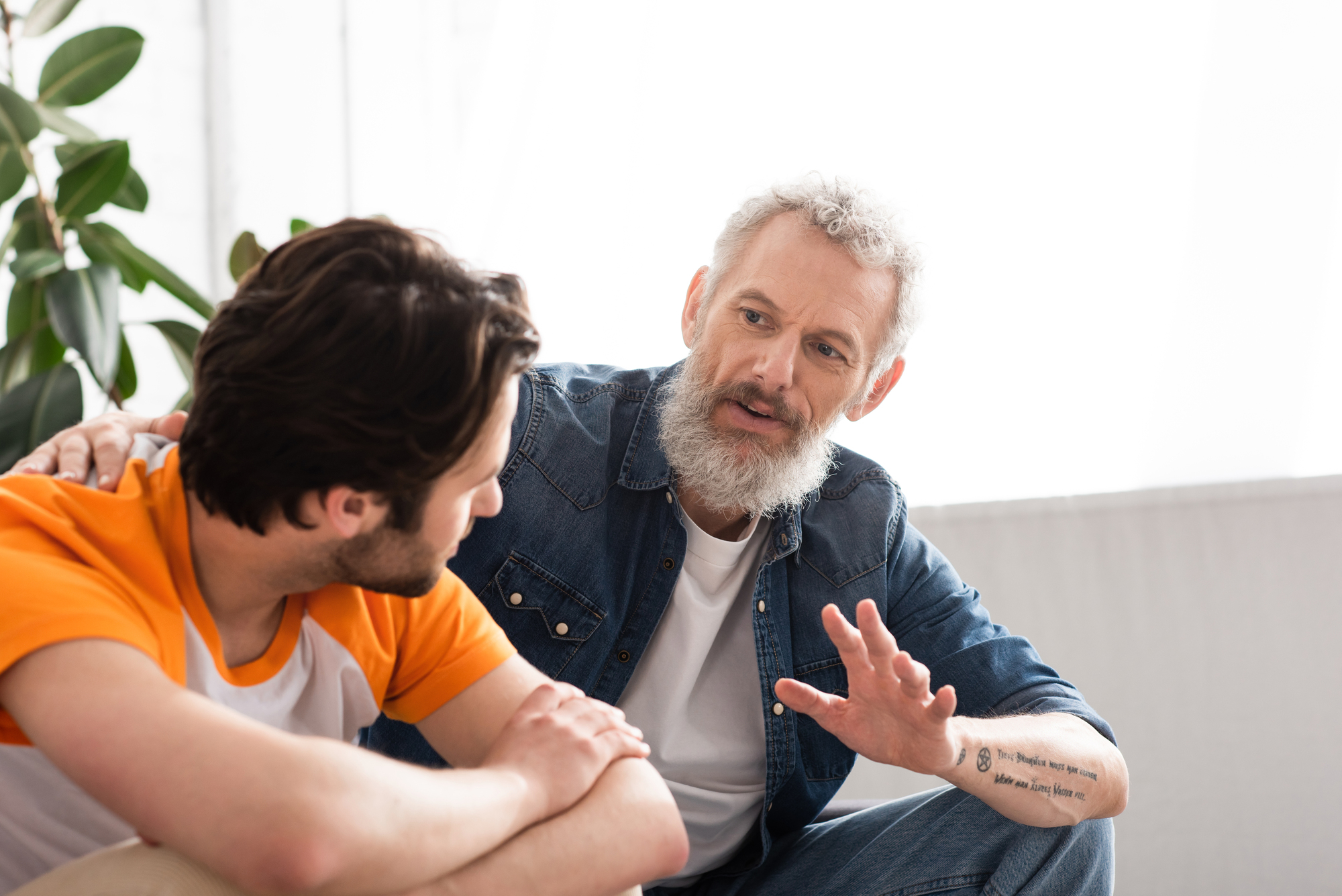 An older man with a grey beard and denim shirt gives advice to a younger man in an orange and white t-shirt. The older man has a tattoo on his forearm. They are sitting in a bright room with a large window and green plants in the background.