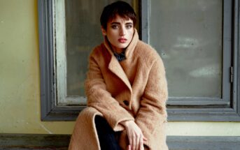 A person with short hair wearing a long, tan coat sits in front of a large window. They have a thoughtful expression and rest one hand on their knee. The window frames a muted, neutral backdrop.