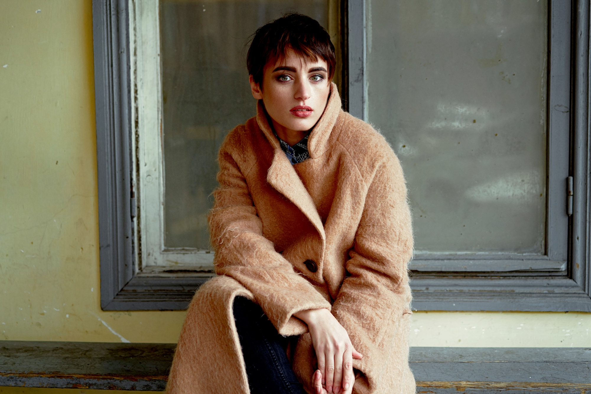 A person with short hair wearing a long, tan coat sits in front of a large window. They have a thoughtful expression and rest one hand on their knee. The window frames a muted, neutral backdrop.