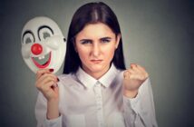 A woman wearing a white shirt holds a happy clown mask in one hand, while her face shows an angry expression. Her other hand is clenched into a fist. The background is a plain, textured gray.