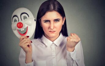 A woman wearing a white shirt holds a happy clown mask in one hand, while her face shows an angry expression. Her other hand is clenched into a fist. The background is a plain, textured gray.