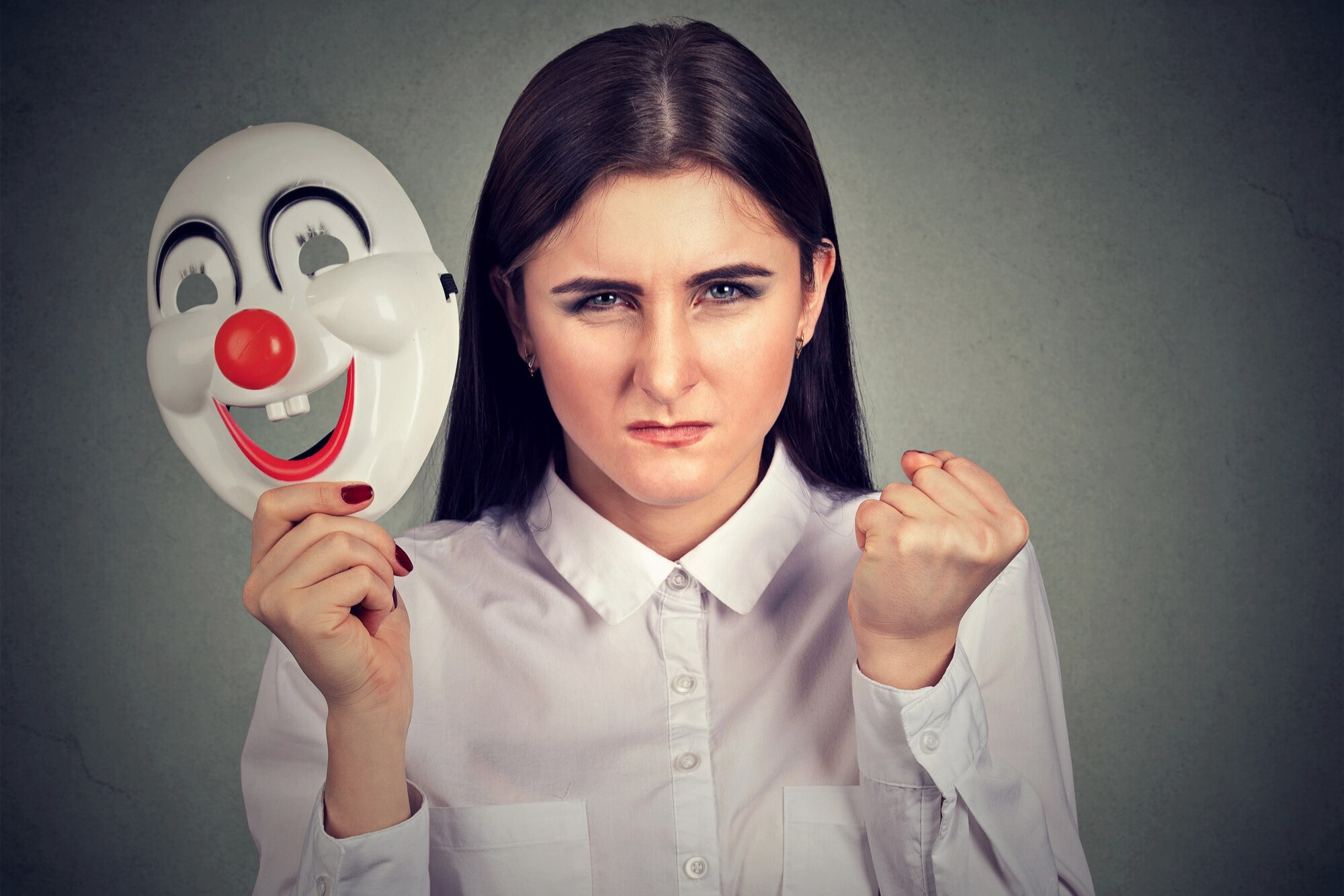 A woman wearing a white shirt holds a happy clown mask in one hand, while her face shows an angry expression. Her other hand is clenched into a fist. The background is a plain, textured gray.