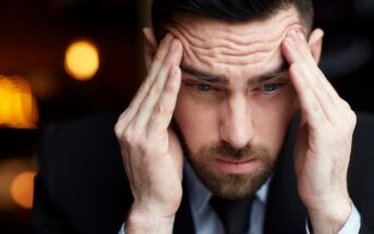 A man in a suit appears stressed, holding both hands to his temples. His eyebrows are furrowed, and he looks down, as if lost in thought or concern. The background is softly blurred with warm lights.