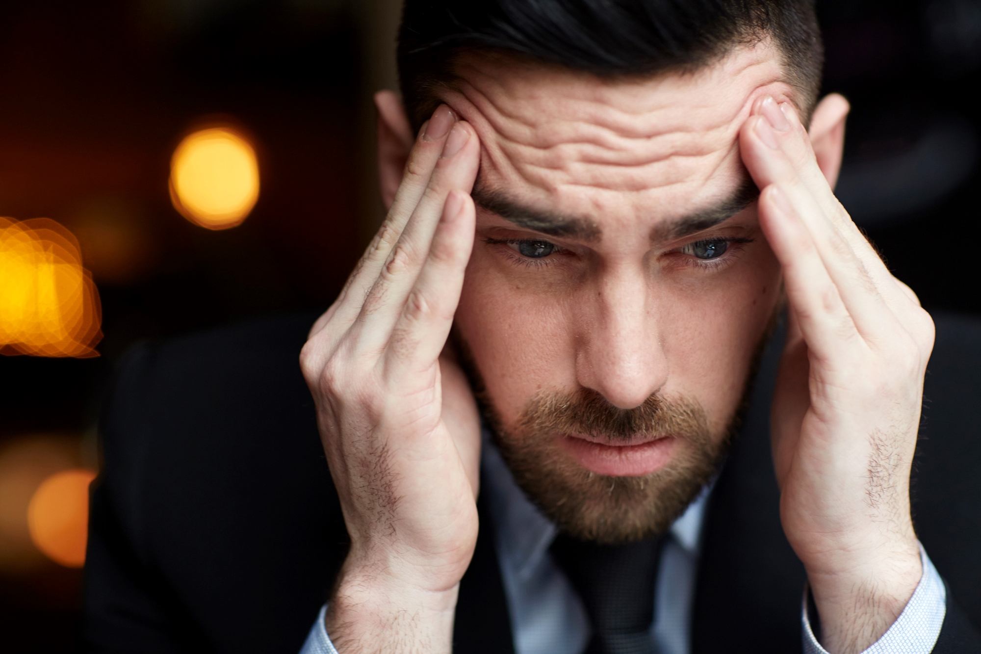 A man in a suit appears stressed, holding both hands to his temples. His eyebrows are furrowed, and he looks down, as if lost in thought or concern. The background is softly blurred with warm lights.