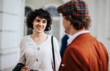 A woman with curly hair in a white blouse is smiling and engaging in conversation with a man wearing a plaid cap and a brown jacket. They are standing indoors near a window.