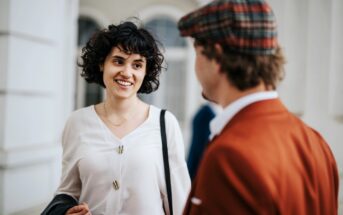 A woman with curly hair in a white blouse is smiling and engaging in conversation with a man wearing a plaid cap and a brown jacket. They are standing indoors near a window.