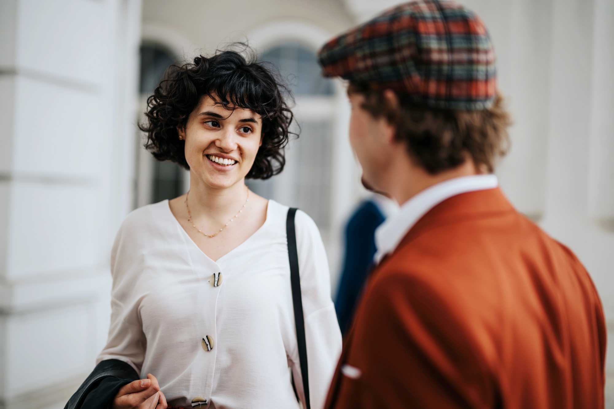 A woman with curly hair in a white blouse is smiling and engaging in conversation with a man wearing a plaid cap and a brown jacket. They are standing indoors near a window.