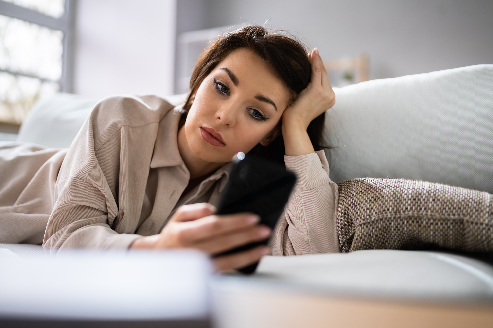 A woman with brown hair and a neutral expression lies on a couch, propping up her head with one hand. She is wearing a beige blouse and looking at her smartphone. Pillows are behind her, and soft light filters through a window.