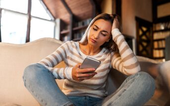 A woman sitting on a sofa, wearing a striped sweater and jeans, looks at her phone with a thoughtful expression, inside a cozy room with a large window and bookshelves.