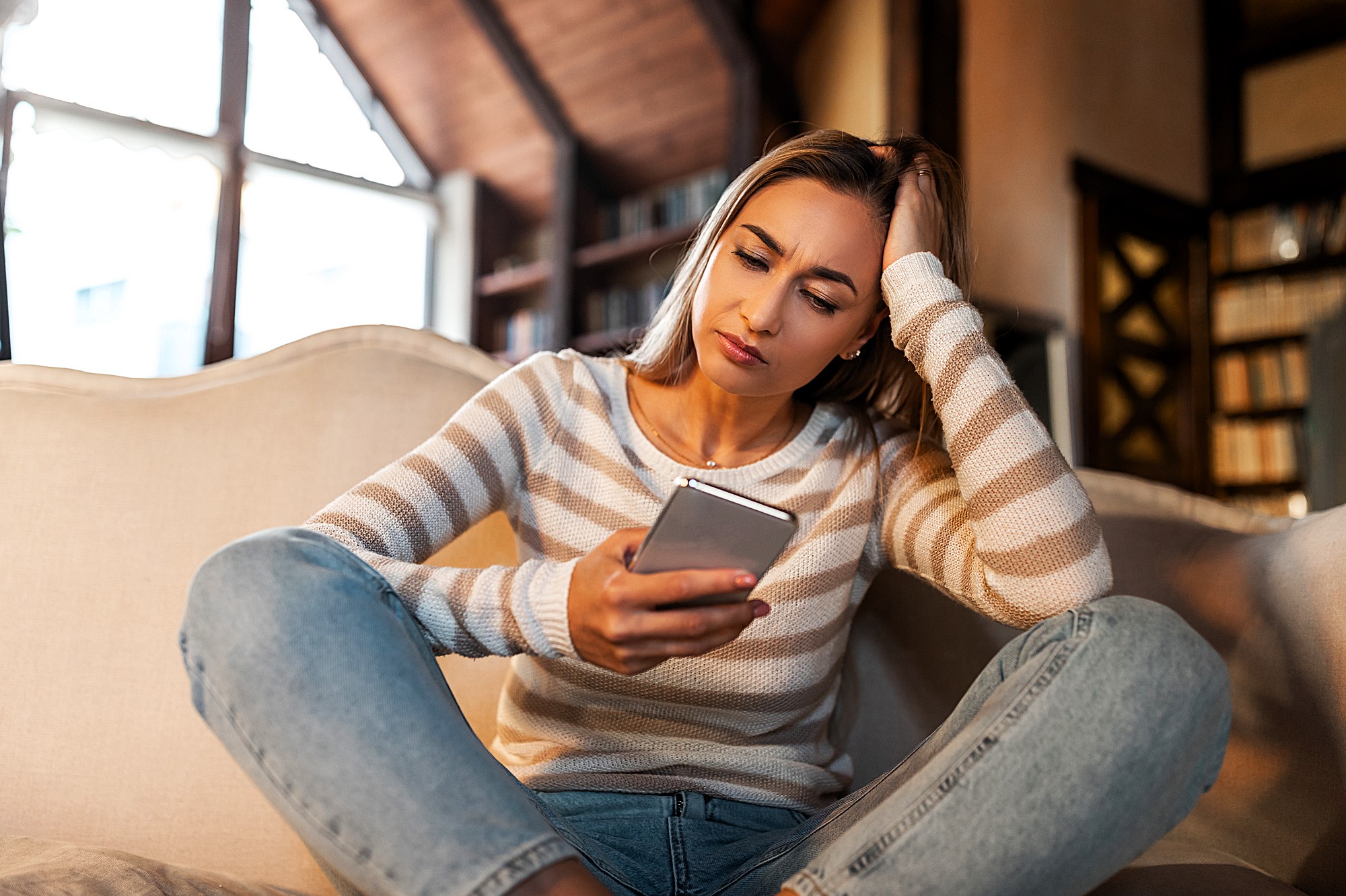 A woman sitting on a sofa, wearing a striped sweater and jeans, looks at her phone with a thoughtful expression, inside a cozy room with a large window and bookshelves.