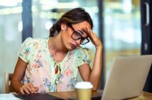 A woman with glasses, wearing a light floral blouse, sits at a table looking stressed in front of a laptop. She holds her forehead with one hand, with a tablet and a coffee cup nearby. The background shows a modern office setting.