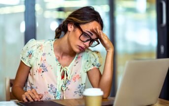 A woman with glasses, wearing a light floral blouse, sits at a table looking stressed in front of a laptop. She holds her forehead with one hand, with a tablet and a coffee cup nearby. The background shows a modern office setting.