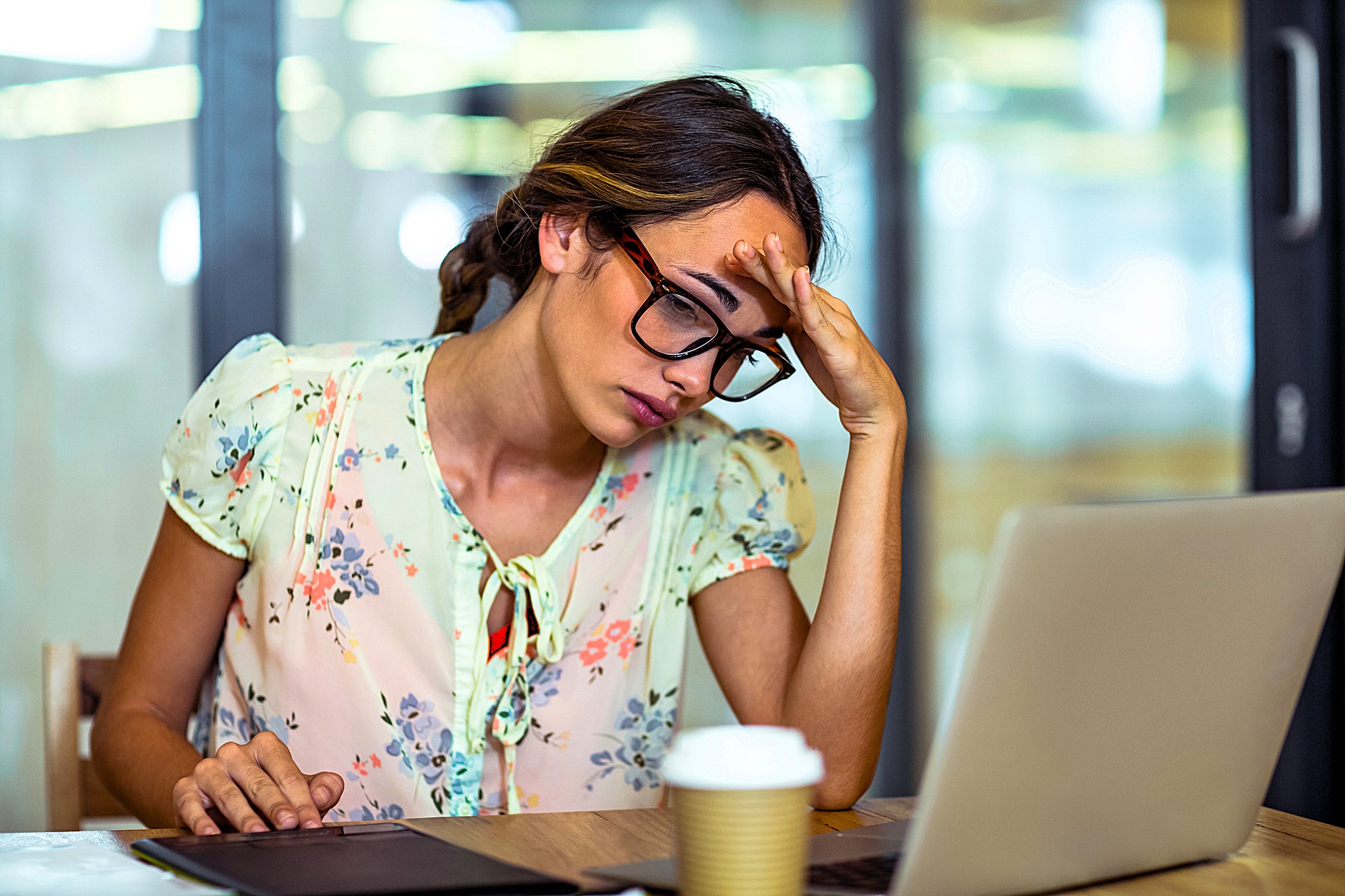 A woman with glasses, wearing a light floral blouse, sits at a table looking stressed in front of a laptop. She holds her forehead with one hand, with a tablet and a coffee cup nearby. The background shows a modern office setting.