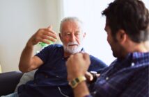 An older man with gray hair and a beard, wearing a blue shirt, is gesturing while talking to a younger man with dark hair, wearing a plaid shirt and a smartwatch, as they sit on a beige sofa in a well-lit room.