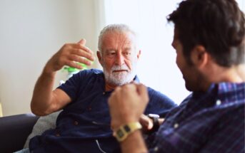 An older man with gray hair and a beard, wearing a blue shirt, is gesturing while talking to a younger man with dark hair, wearing a plaid shirt and a smartwatch, as they sit on a beige sofa in a well-lit room.