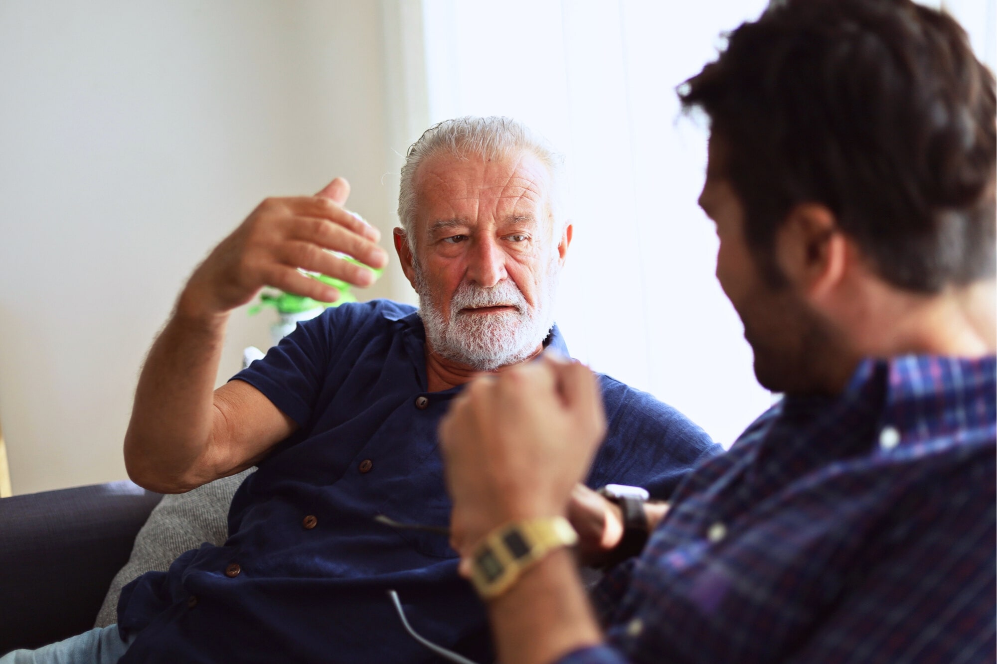 An older man with gray hair and a beard, wearing a blue shirt, is gesturing while talking to a younger man with dark hair, wearing a plaid shirt and a smartwatch, as they sit on a beige sofa in a well-lit room.