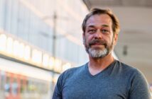 A man with a beard and mustache is standing outdoors, wearing a gray T-shirt. The background is slightly blurred, showing a modern building with glass windows and soft evening lighting.