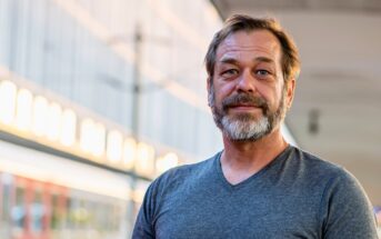 A man with a beard and mustache is standing outdoors, wearing a gray T-shirt. The background is slightly blurred, showing a modern building with glass windows and soft evening lighting.