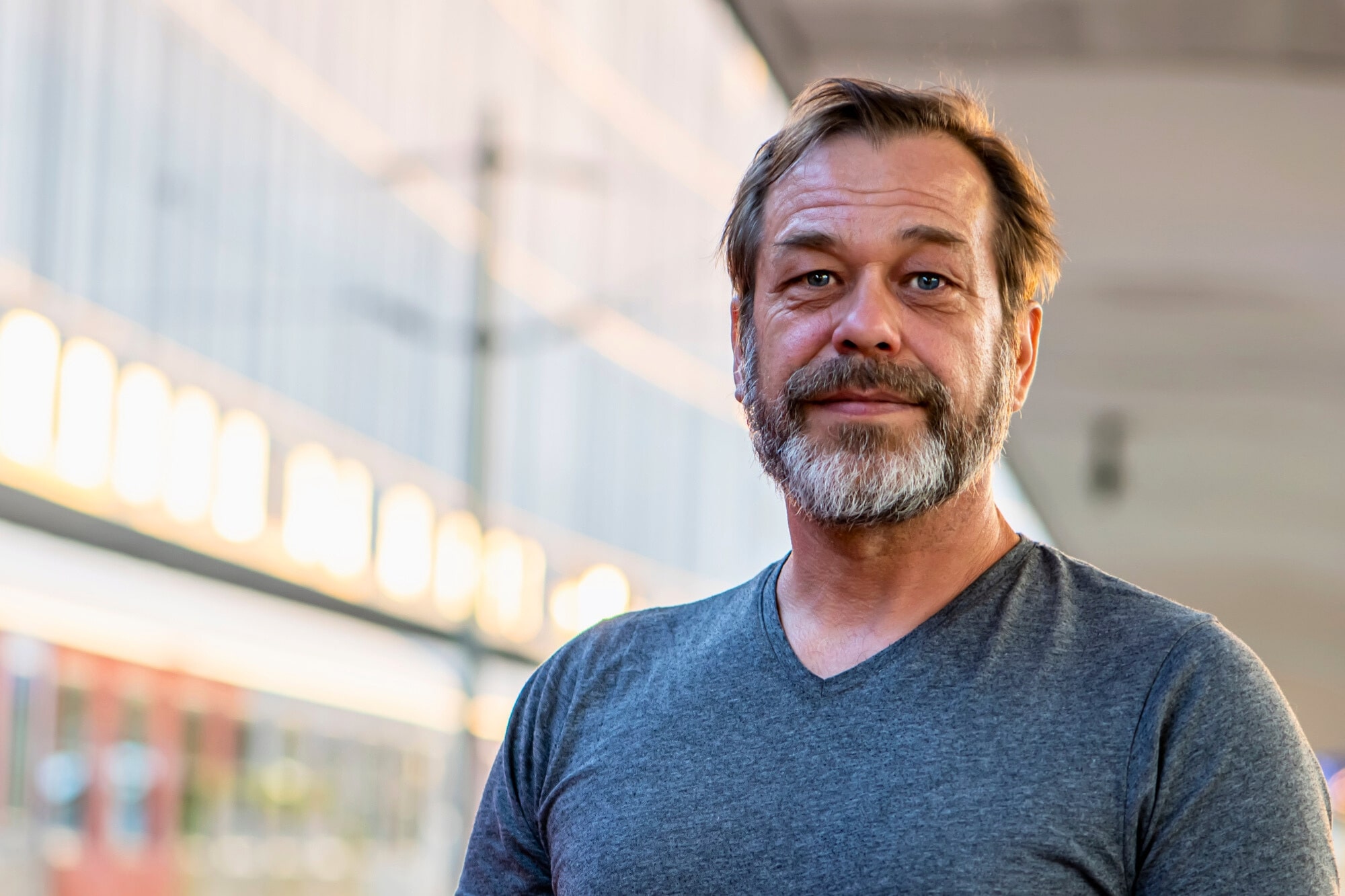 A man with a beard and mustache is standing outdoors, wearing a gray T-shirt. The background is slightly blurred, showing a modern building with glass windows and soft evening lighting.