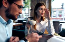 A woman in a white shirt holds a coffee cup, looking to the side thoughtfully. A man in glasses sits next to her, using a smartphone. They are in a cafe with large windows and wooden tables. Papers are spread out on the table.