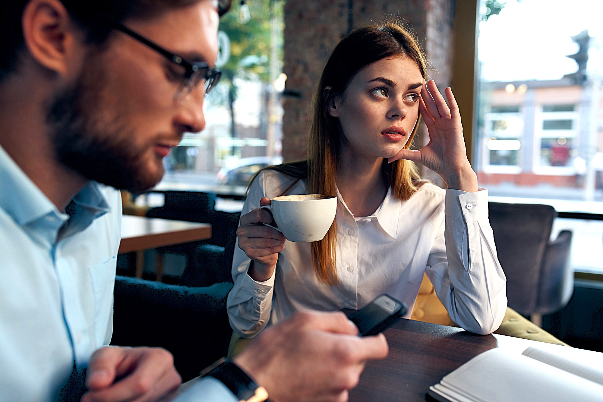 A woman in a white shirt holds a coffee cup, looking to the side thoughtfully. A man in glasses sits next to her, using a smartphone. They are in a cafe with large windows and wooden tables. Papers are spread out on the table.