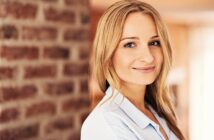 Smiling woman with long blonde hair wearing a light blue shirt stands in front of a brick wall. The background is softly blurred, highlighting her cheerful expression.