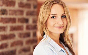 Smiling woman with long blonde hair wearing a light blue shirt stands in front of a brick wall. The background is softly blurred, highlighting her cheerful expression.