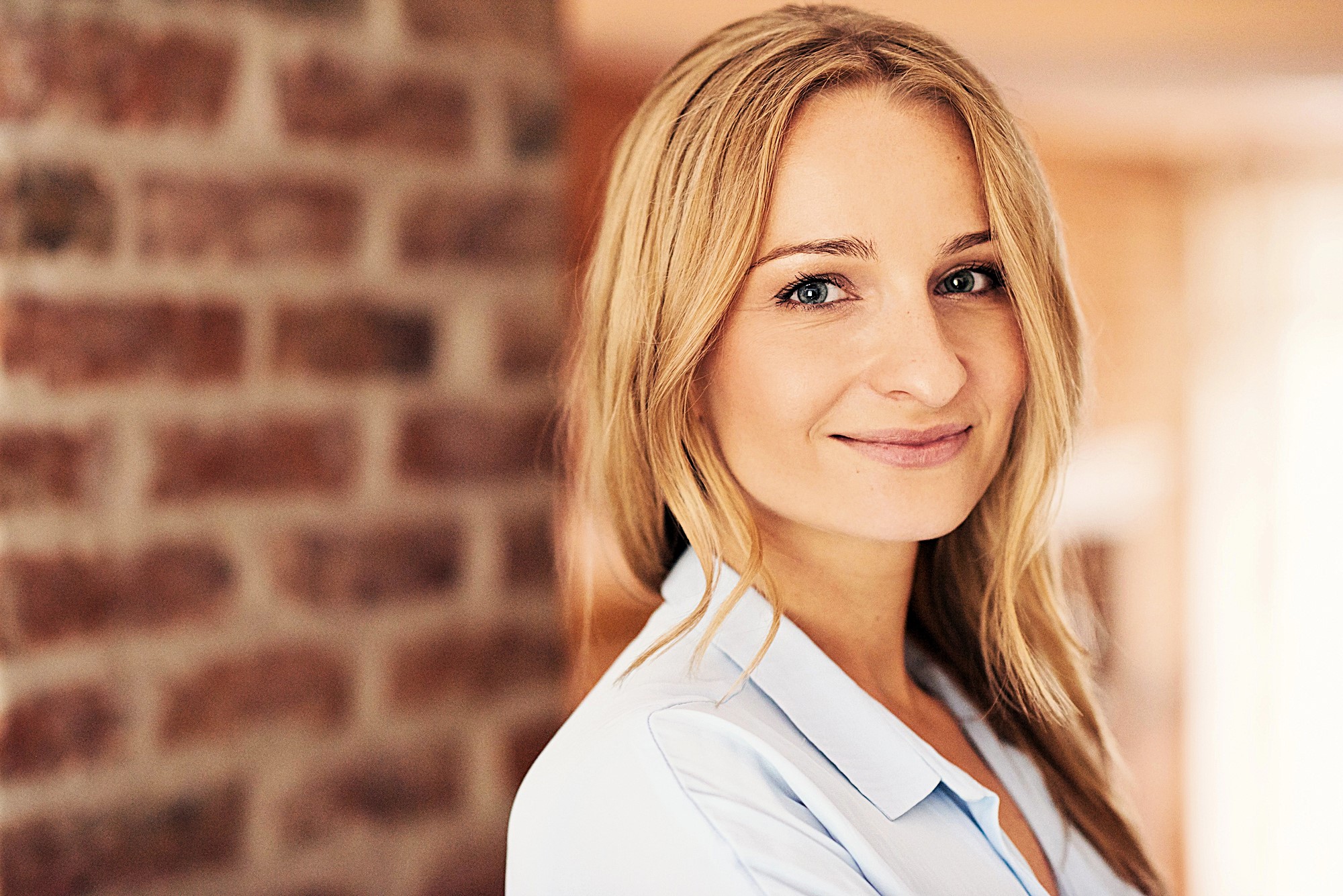 Smiling woman with long blonde hair wearing a light blue shirt stands in front of a brick wall. The background is softly blurred, highlighting her cheerful expression.