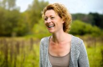 A woman with curly blonde hair smiles joyfully while standing in a lush, green field. She is wearing a gray cardigan and a beige top. The background features trees and a slightly overcast sky, creating a serene, natural setting.