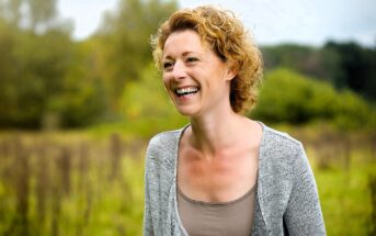 A woman with curly blonde hair smiles joyfully while standing in a lush, green field. She is wearing a gray cardigan and a beige top. The background features trees and a slightly overcast sky, creating a serene, natural setting.