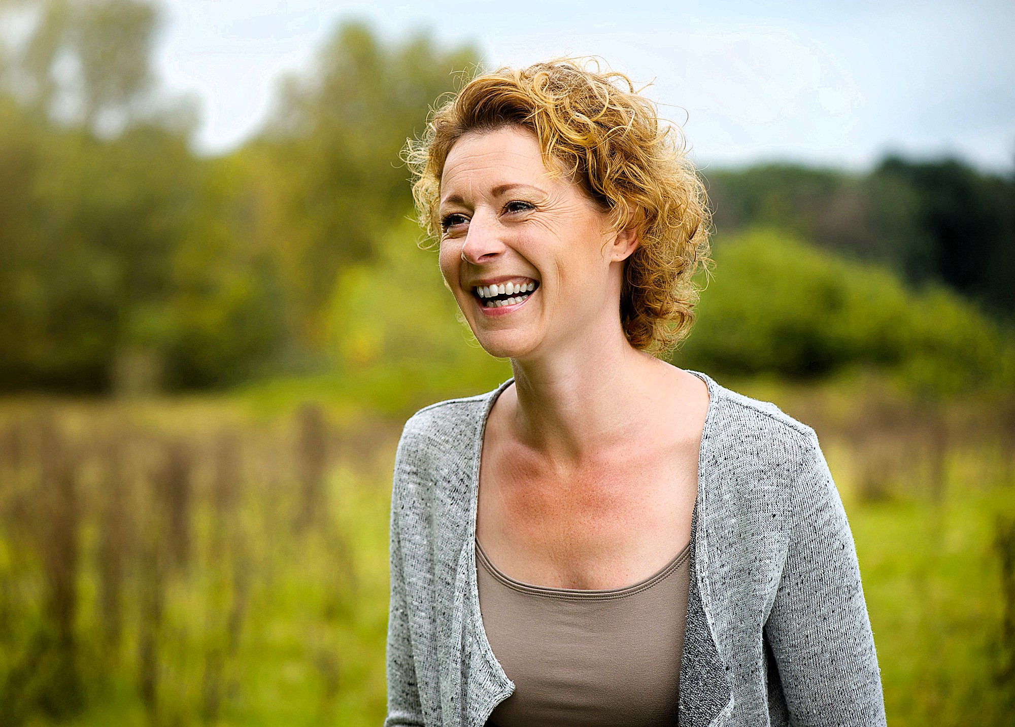 A woman with curly blonde hair smiles joyfully while standing in a lush, green field. She is wearing a gray cardigan and a beige top. The background features trees and a slightly overcast sky, creating a serene, natural setting.