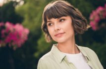 A woman with short brown hair gazes to the side, standing outdoors. She is wearing a light green shirt over a white top. There are vibrant pink flowers and lush greenery in the blurred background.