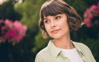 A woman with short brown hair gazes to the side, standing outdoors. She is wearing a light green shirt over a white top. There are vibrant pink flowers and lush greenery in the blurred background.