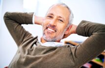 A smiling man with gray hair and a beard, wearing a brown sweater over a white shirt, leans back in his chair with his hands behind his head, appearing relaxed and content.