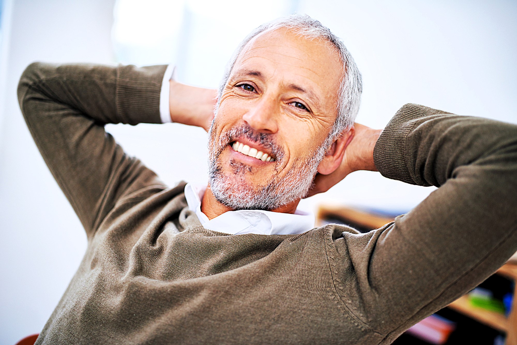 A smiling man with gray hair and a beard, wearing a brown sweater over a white shirt, leans back in his chair with his hands behind his head, appearing relaxed and content.