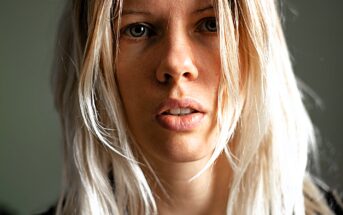 A close-up of a person with long, light blonde hair, looking directly at the camera. The background is blurred, focusing attention on the person's face and expression. The lighting highlights natural features and textures.