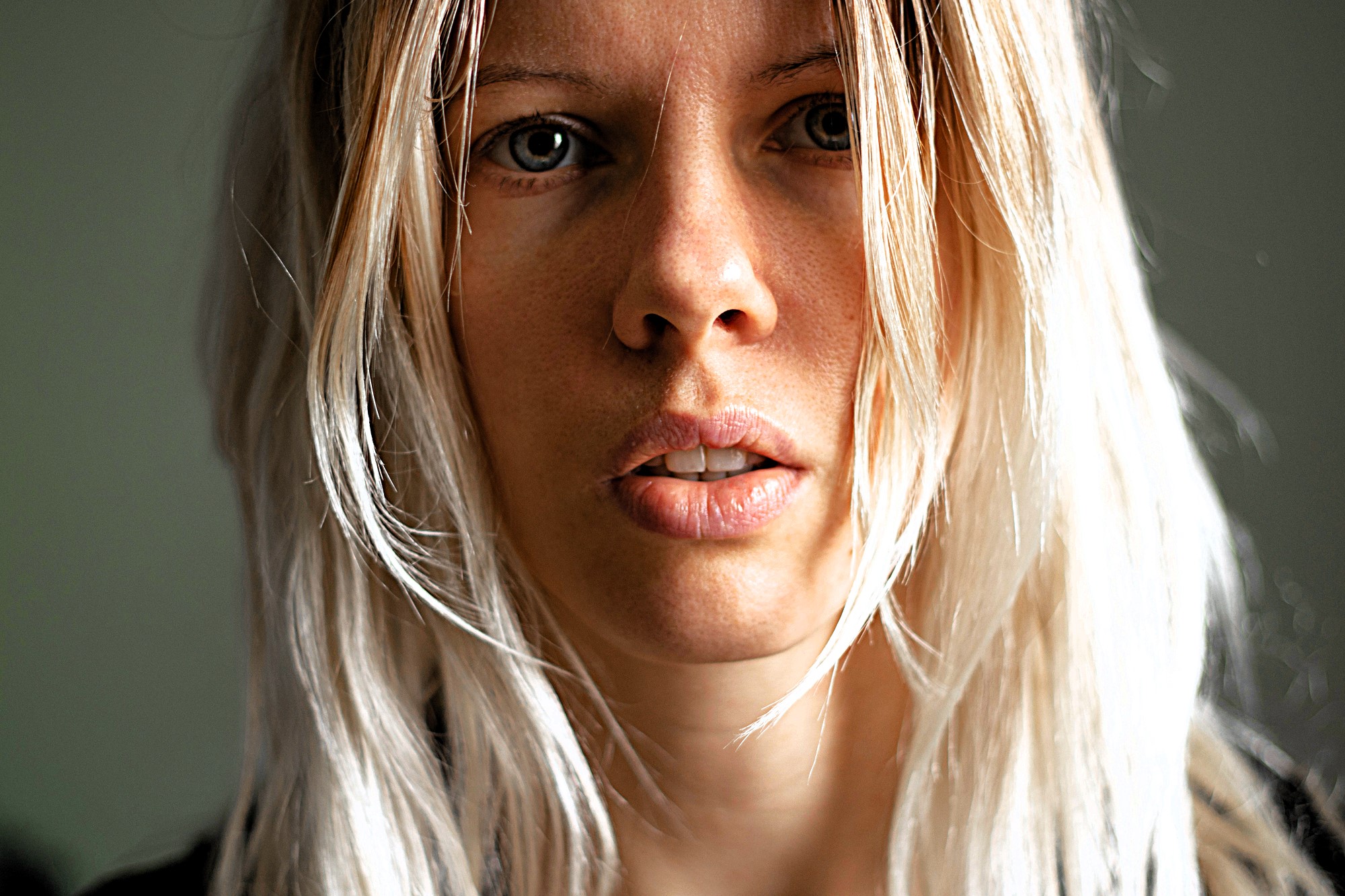 A close-up of a person with long, light blonde hair, looking directly at the camera. The background is blurred, focusing attention on the person's face and expression. The lighting highlights natural features and textures.