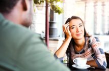 A woman sits at an outdoor cafe table with a cup of coffee, resting her head on her hand. She gazes thoughtfully at a person across from her, who is partially visible. The background features potted plants and a blurred street view.