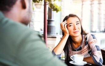 A woman sits at an outdoor cafe table with a cup of coffee, resting her head on her hand. She gazes thoughtfully at a person across from her, who is partially visible. The background features potted plants and a blurred street view.