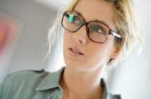 A person with light hair wearing glasses looks off to the side. They are dressed in a light-colored button-up shirt, and the background is softly blurred.