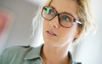 A person with light hair wearing glasses looks off to the side. They are dressed in a light-colored button-up shirt, and the background is softly blurred.