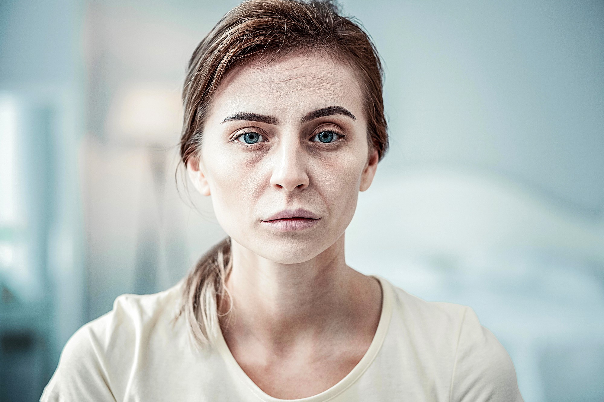 A woman with light brown hair tied back, wearing a beige shirt, stares directly at the camera with a neutral expression. The background is softly blurred, suggesting an indoor setting.