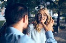 A woman with glasses smiles while talking to a man, sitting outdoors on a bench in a park. She is listening attentively, and there is a blurred background of trees and pathways.