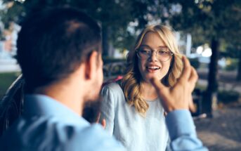 A woman with glasses smiles while talking to a man, sitting outdoors on a bench in a park. She is listening attentively, and there is a blurred background of trees and pathways.