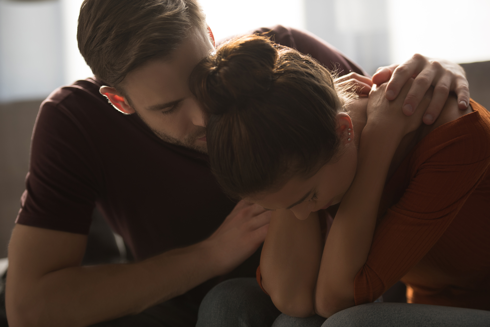 A man gently comforts a woman who appears upset. She sits with her head bowed and arms folded, while he places a reassuring hand on her shoulder and leans close. The mood is calm and supportive, and the setting seems to be indoors.