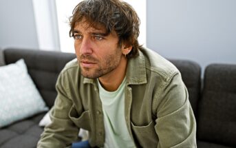 A man with brown hair and a beard sits on a gray couch, wearing a green button-up shirt over a white T-shirt. He appears thoughtful, looking slightly upwards. Pillows are visible on the couch. The room has soft natural lighting.