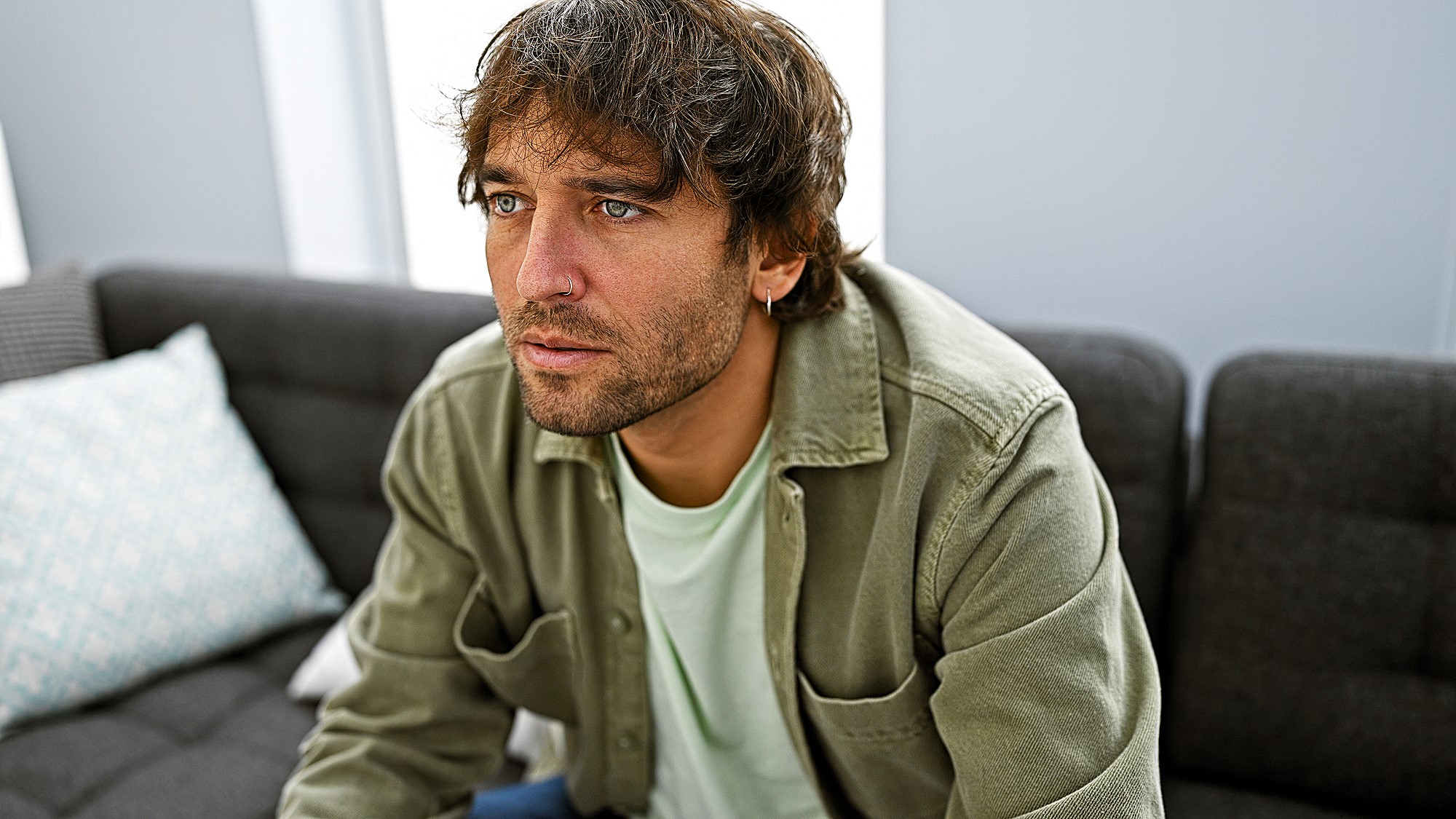 A man with brown hair and a beard sits on a gray couch, wearing a green button-up shirt over a white T-shirt. He appears thoughtful, looking slightly upwards. Pillows are visible on the couch. The room has soft natural lighting.